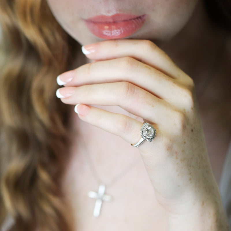 Close-up of a woman's hand with a diamond ring, wearing a white dress with a blurred background.