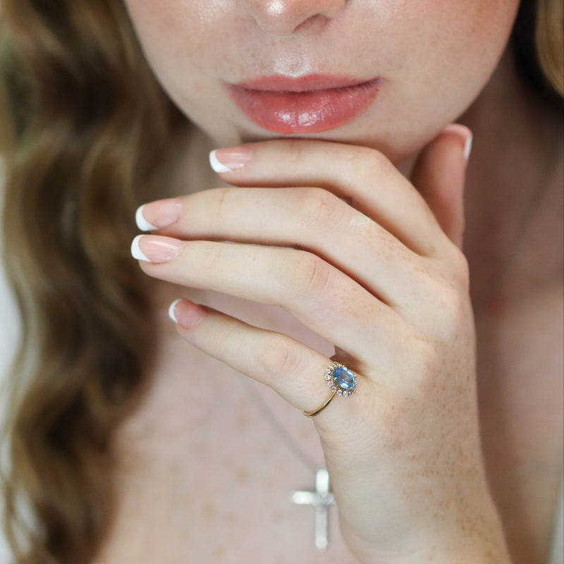 Close-up of a woman's hand wearing a ring with a diamond and blue topaz, against a blurred background.